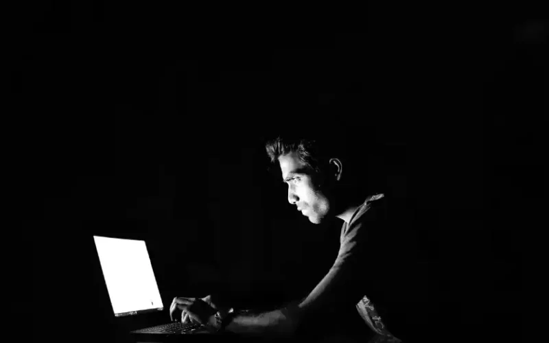 Eine düstere Szene: Ein junger Mann sitzt vor einem Notebook und wird nur vom hellen Licht des Bildschirms beschienen. Man kann nicht erkennen, was auf dem Laptop dargestellt wird.